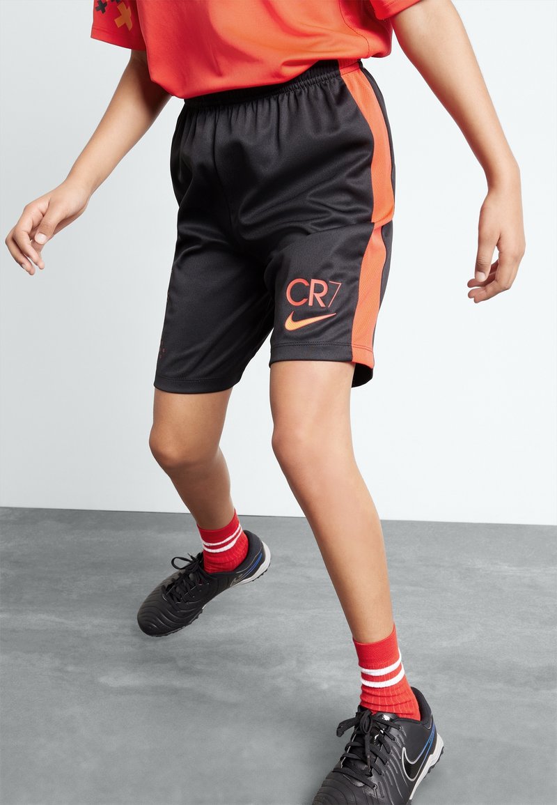 Niño que lleva puestos pantalones cortos deportivos CR7 en negro y naranja, calcetines rojos con rayas blancas, botas Nike negras y una camiseta deportiva roja, de pie sobre un suelo gris.