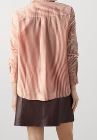Person wearing long-sleeve red and white striped shirt and dark brown leather skirt, standing against a plain light background, facing away.