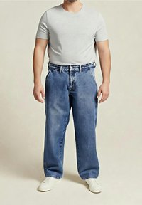 Homme portant un t-shirt gris clair uni, un jean bleu ample et des baskets blanches, debout devant un fond uni.