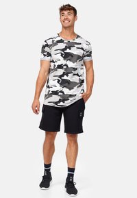 T-shirt camouflage gris à manches courtes, associé à un short cargo noir. Le modèle se tient de façon décontractée avec une main dans la poche.