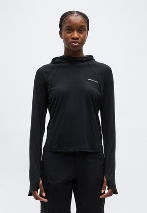 Γυναίκα με μαύρο μακρυμάνικο φούτερ Columbia και μαύρο παντελόνι, στέκεται μπροστά σε απλό ανοιχτό γκρι φόντο, κοιτάζοντας προς τα εμπρός.