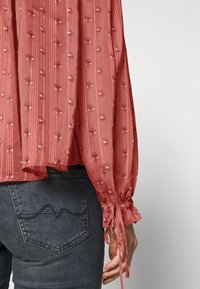 Blusa de coral claro con patrón texturizado, de mangas largas, con puños fruncidos y lazos, y un ajuste holgado, combinada con jeans de mezclilla oscura.