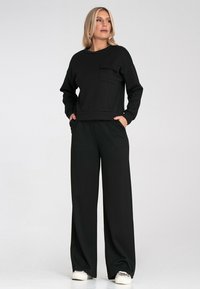 Czarny bawełniany sweatshirt z okrągłym dekoltem i kieszenią, w poł�ączeniu z szerokimi czarnymi spodniami. Model stoi z rękami w kieszeniach, nosząc trampki.