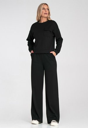 Czarny bawełniany sweatshirt z okrągłym dekoltem i kieszenią, w połączeniu z szerokimi czarnymi spodniami. Model stoi z rękami w kieszeniach, nosząc trampki.