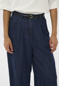 Oseba nosi temno modre kavbojke visokega pasu z črnim pasom in belim kratkim majico, ki je vtaknjena, roke sproščene ob straneh.