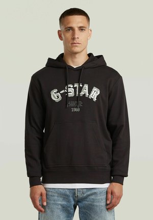 Ung mand iført sort G-Star hættetrøje med lomme foran og lyseblå jeans, stående mod en ensfarvet lys baggrund.