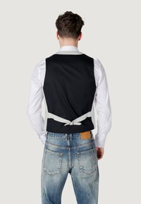 Gilet nero con una cravatta a righe in vita, indossato sopra una camicia bianca, abbinato a jeans azzurri chiari con una toppa di pelle sul retro.