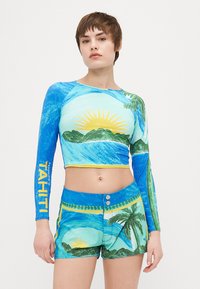 Långärmad crop top i blått med en tryckt sol, hav och palmer; kombinerad med matchande shorts med liknande tropiska mönster.