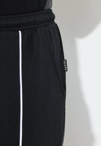 Czarne sportowe spodnie z regulowanym paskiem ze sznurkiem, boczną kieszenią i białym paskiem wzdłuż zewnętrznego szwu. Miękka tkanina.