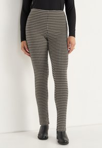 Vrouw in zwart en beige hounds-tooth patroon slim-fit broek met zwarte enkellaarzen en een zwart langemouwen top.