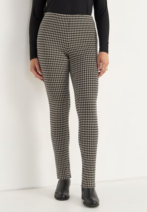 Nainen, joka on pukeutunut mustavalkoiseen houndstooth-kuvioiseen slim-fit-housuun, mustiin nilkkureihin ja mustaan pitkähihaiseen puseroon.