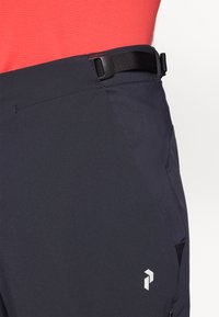 Sorte softshell bukser med et stilrent design, der har bæltestropper, en lynlåslomme og et hvidt logo foran. Glat tekstur.