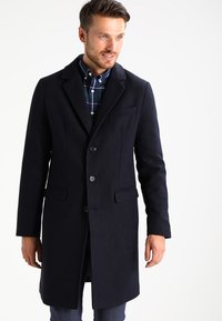 Navy wool blend coat with a tailored fit, notched lapels, front buttons, and side pockets. Worn over a blue checkered shirt.