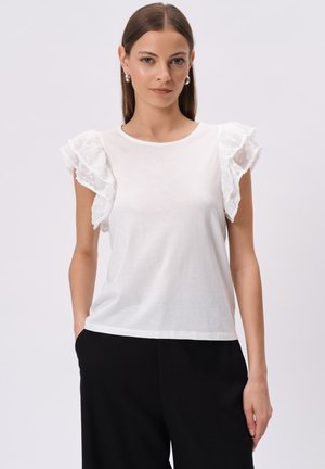 Biała bluzka z krótkim rękawem, z falbaniastego przezroczystego materiału w kropki. Miękka tkanina, okrągły dekolt. Połączona z czarnymi, luźnymi spodniami.