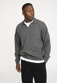 Grå ribbet pullover trøje med krave og lange ærmer, lagd oven på en hvid T-shirt. Sorte bukser fuldender outfittet.