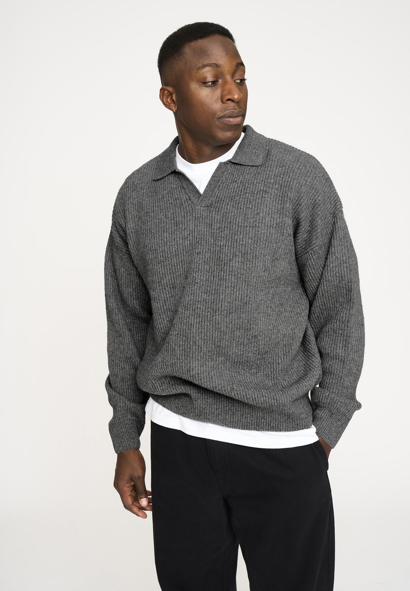 Grå ribbet pullover trøje med krave og lange ærmer, lagd oven på en hvid T-shirt. Sorte bukser fuldender outfittet.