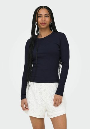 Femme aux longs cheveux tressés portant un haut ajusté à manches longues foncé et un short blanc à motif œillet, debout devant un fond uni.
