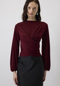 Burgundska dolga majica s kratkimi rokavi z nabranim dizajnom na pasu, v kombinaciji s črno prilegajočo krilom, ki ima gladko teksturo in preprosto silhueto.