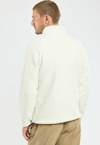 Pullover en polaire blanc, texturé, avec un col montant, des manches longues et une coupe décontractée. Le tissu semble doux et confortable.