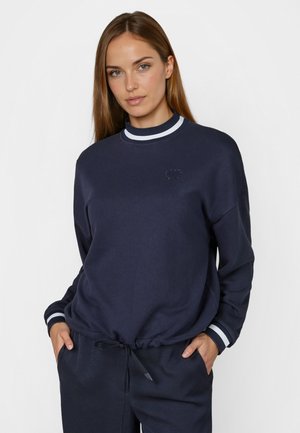 Woman wearing a navy blue sweatshirt and pants with white-striped cuffs, hands in pockets, and a small stitched heart on the chest.