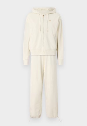 Felpa con zip beige, dotata di lacci e tasche frontali, abbinata a pantaloni jogger affusolati con caviglia ristretta e branding discreto.