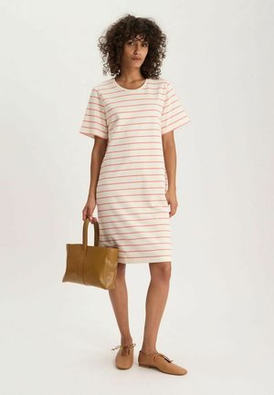 Femme aux cheveux bouclés portant une robe blanche à fines rayures rouges horizontales, tenant un sac à main marron et portant des chaussures à lacets beige.