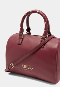 Bordeauxrote Lederhandtasche mit doppelten Griffen, verstellbarem Schulterriemen, Goldfarbenem Beschläge, strukturiertem Finish und geprägtem Logo an der Seite.