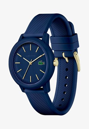 Orologio in silicone blu navy con cinturino testurizzato, indicatori delle ore dorati e logo Lacoste. La cassa rotonda presenta un design minimalista per una visibilità chiara.