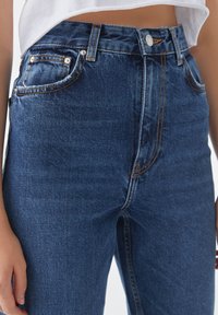 Jeans en denim taille haute de couleur bleu foncé avec des coutures dorées. Comprend des poches, une fermeture éclair et une silhouette ajustée. Le tissu a une texture douce.