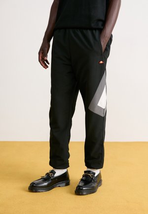 Homme portant un pantalon de survêtement noir avec un motif géométrique gris et blanc sur le côté, des mocassins noirs, des chaussettes blanches, debout sur un sol couleur moutarde.
