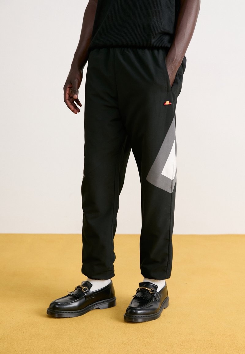 Homme portant un pantalon de survêtement noir avec un motif géométrique gris et blanc sur le côté, des mocassins noirs, des chaussettes blanches, debout sur un sol couleur moutarde.
