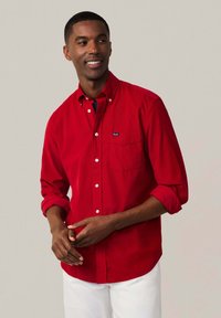 Camisa roja de botones, hecha de algodón, con mangas largas, puños enrollables y un bolsillo frontal. Presenta una pequeña etiqueta con un logo en el pecho.