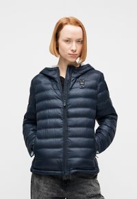 Νέος με κοντά κόκκινα μαλλιά που φοράει μπλε σκούρο καπιτονέ μπουφάν, με τα χέρια στις τσέπες, στέκεται μπροστά σε απλό λευκό φόντο.