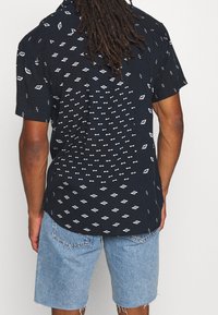 Kortärmad svart skjorta med vita geometriska mönster, med krage som kan knappas ner och en avslappnad passform. Bärs med ljusa jeansshorts.