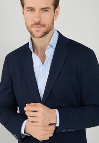 Blazer navy slim fit, realizzato in tessuto testurizzato. Indossato sopra una camicia a righe azzurre, con bottoni bianchi e polsini a contrasto.