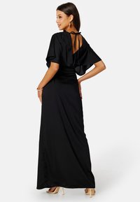 Abito maxi nero con maniche a sbuffo, schiena scoperta e dettaglio con fiocco; texture liscia e silhouette aderente che accentua le curve. Tacchi alti visibili.