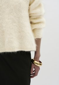 Romige, fluffy gebreide trui met losse mouwen; gedragen over een strakke zwarte rok. De pols van het model heeft een glanzende, gouden bangle armband.