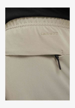 Beige DRI-FIT bukser med elastisk linning, sidelommer med glidelås og jevn tekstur. Sømmen er tonet og minimal.