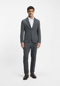 Completo grigio su misura realizzato in tessuto testurizzato, caratterizzato da un blazer con un solo bottone, due tasche frontali e pantaloni abbinati, indossato con una camicia bianca.