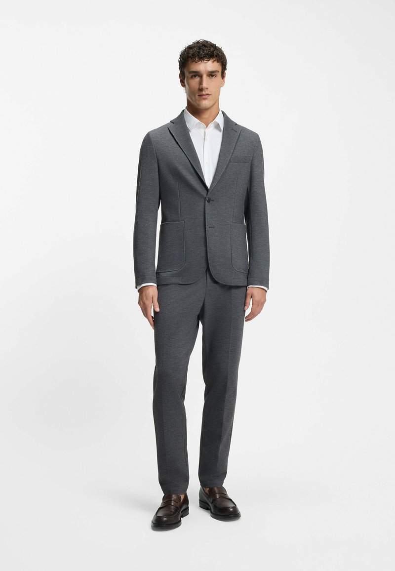 Completo grigio su misura realizzato in tessuto testurizzato, caratterizzato da un blazer con un solo bottone, due tasche frontali e pantaloni abbinati, indossato con una camicia bianca.