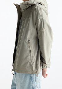 Veste légère, verte olive, avec capuche, poches latérales zippées, poignets élastiques et un logo circulaire sur la manche. Texture de tissu lisse.