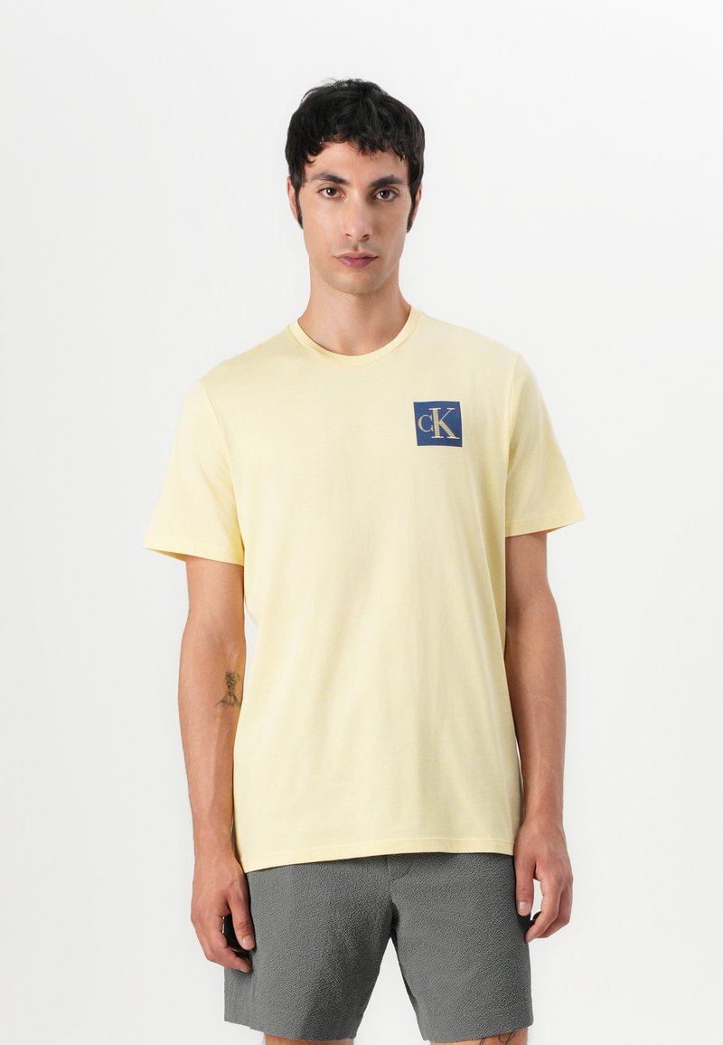 Camiseta de algodón amarilla de manga corta con un logo cuadrado azul que lleva "cK" en blanco. Diseño sencillo con cuello redondo y corte relajado.