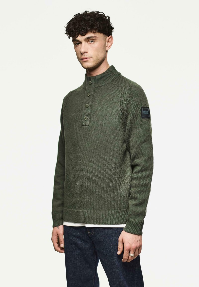Mann trägt einen geknöpften olivgrünen Strickpullover mit hohem Kragen und einem Patch an der linken Ärmel, kombiniert mit dunkelblauen Jeans.