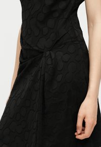 Robe noire avec une texture subtile de léopard en relief et un détail froncé à la taille, montrée sur le torse et le bras d'une personne.