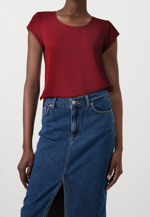 Femme portant un haut rouge à manches courtes ajusté et une jupe en jean bleue taille haute avec une fente frontale sur un fond clair uni.