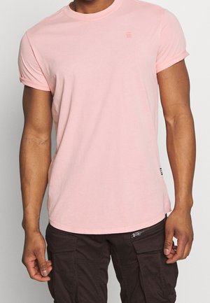 Mann trägt ein schlichtes hellrosa kurzärmeliges T-Shirt und dunkelbraune Hose mit Reißverschlusstasche, steht vor einem schlichten hellen Hintergrund.
