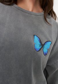Close-up of a gray shirt with a bright blue butterfly print on the chest and visible neckline and hair strands.