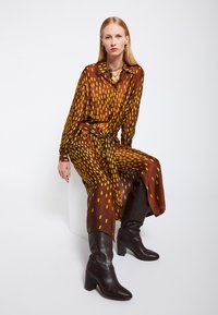 Abito marrone a maniche lunghe con un motivo geometrico giallo, dotato di una vita cintata e colletto. Abbinato a stivali marroni con tacco alti fino al ginocchio.