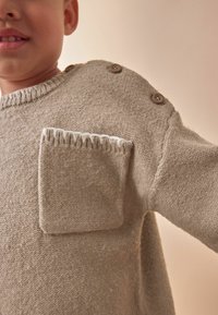 Beige neulepaita pyöreällä kauluksella, jossa on neliön muotoinen tasku ja kaksi ruskeaa nappia olkapäällä. Pehmeä tekstiili ja rento muotoilu.