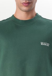 Sweatshirt verde închis din bumbac, cu guler rotund și un mic logo alb care spune "SELECTED" pe partea stângă a pieptului.